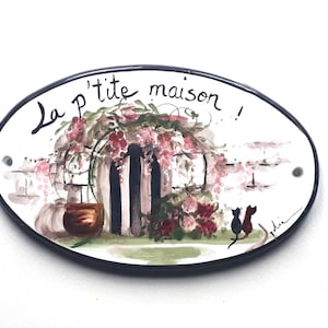 Puede incluir: Un letrero ovalado de cerámica blanca con borde negro. El letrero tiene una imagen pintada de una casa con una puerta y flores. El texto "La p'tite maison!" está escrito en cursiva negra en el letrero.