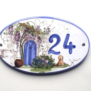 以下が含まれることがあります： 青いドア、フジの花、茶色の鉢、ゴールデンレトリバーの犬が描かれた、楕円形のセラミック製の家の番号札。番号の24は青で書かれている。