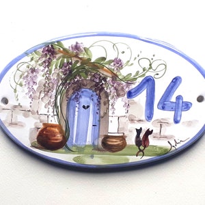 May include: Oval ceramic house number sign featuring a blue door, wisteria vines, and two cats. The number 14 is painted in blue on the sign.