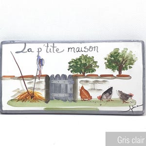 Puede incluir: Un azulejo de cerámica blanco con una escena pintada a mano de una casita con una valla, dos árboles y gallinas. El azulejo tiene el texto "La p'tite maison" escrito en pintura negra.