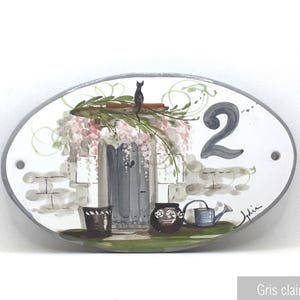 May include: White oval ceramic house number sign with a grey border. The number 2 is painted in grey. The sign features a painted scene of a grey door with a black cat sitting on top, a brick wall, and a potted plant. The sign is decorated with a floral design.
