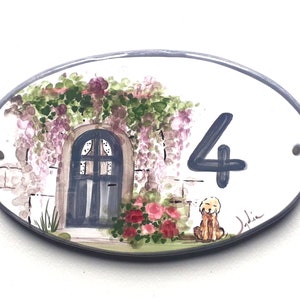 May include: Oval ceramic house number sign featuring a painted design of a door with a vine covered archway, a dog sitting in front of the door, and the number 4 in blue paint. The sign is painted on a white background with a light grey border.