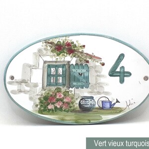 May include: Oval ceramic house number sign featuring a painted scene of a window with green shutters, flowers, and a watering can. The number 4 is painted in turquoise on the sign.
