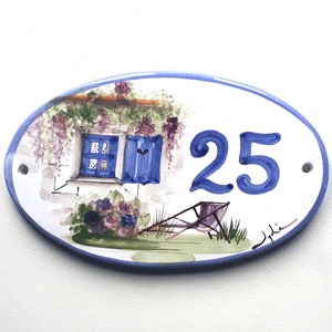 May include: Oval ceramic house number sign with a blue and white painted design of a house with a window and a chair in the yard. The number 25 is painted in blue on the sign.