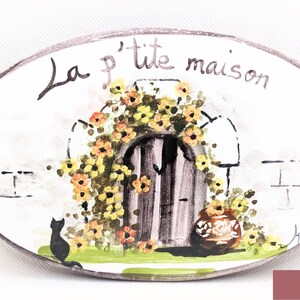 May include: A white oval ceramic sign with a hand-painted design of a small house with a door and flowers. The sign says "La p'tite maison" in French. There is a black cat in the bottom left corner of the sign.