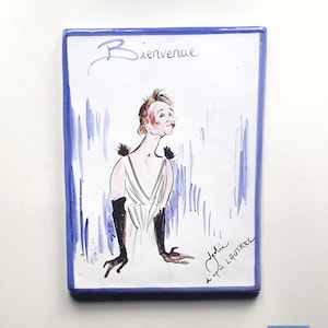 May include: A blue and white ceramic tile featuring a cartoon illustration of a woman in a white dress and black gloves. The tile includes the text "Bienvenue" and "d'apres L'AUTREC" in cursive writing.