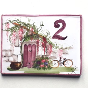 May include: A hand-painted ceramic house number tile with a pink border. The tile features a whimsical illustration of a pink door with a vine of purple flowers, a brick wall, a brown pot, and a bicycle. The number "2" is painted in pink on the tile.