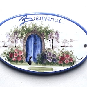 May include: Oval ceramic sign with a blue door, flowers, and a black cat. The sign says "Bienvenue" in cursive.