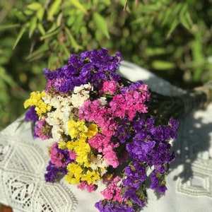 May include: A colourful bouquet of dried flowers in shades of purple, pink, yellow, and white. The flowers are tied with twine and rest on a white lace tablecloth. The background is blurred green foliage.