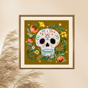 May include: A framed art print featuring a white skull decorated with floral and leaf patterns. The skull is surrounded by a green snake, orange flowers, yellow lilies, and stars against a dark olive background. The frame is gold.