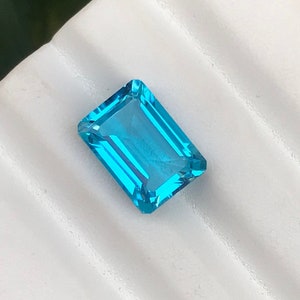 May include: A vibrant blue gemstone with a rectangular emerald cut. The gemstone is displayed on a white surface.