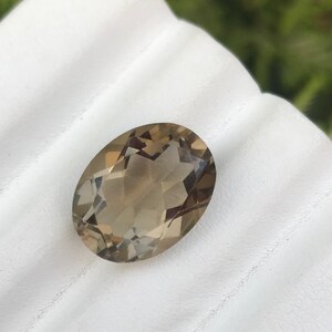 May include: A light brown oval-shaped gemstone with facets. The gemstone is on a white surface.
