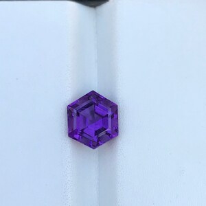May include: A purple gemstone with a hexagonal shape, displayed in a black and white box.