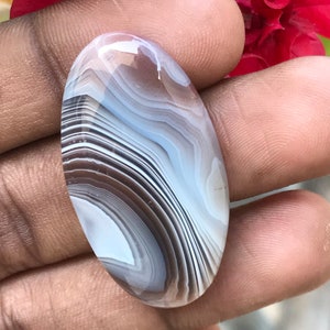 May include: A white and brown agate cabochon with a banded pattern. The cabochon is oval shaped and has a smooth, polished surface.