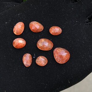May include: Eight orange sunstone cabochons with a sparkly, iridescent finish. The stones are oval shaped and have a smooth, polished surface.