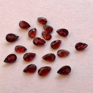 May include: A collection of 17 pear-shaped red gemstones, likely garnets, scattered on a white surface.