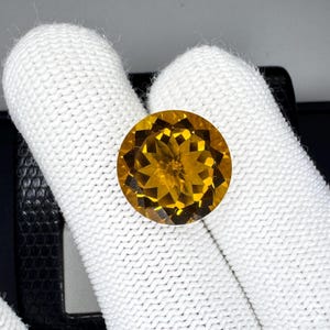 May include: A round, faceted gemstone with a warm, golden-yellow hue. The gem is held against a white background, showcasing its brilliant cut and clarity. The gemstone's facets reflect light, creating a sparkling effect. This gemstone is suitable for jewelry.