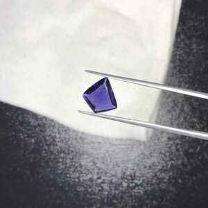 May include: A deep purple gemstone with a faceted, triangular shape, held by tweezers against a white background.