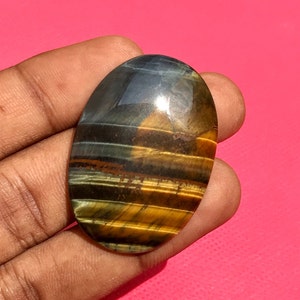 May include: Oval-shaped gemstone with a tiger's eye pattern in shades of brown, gold, and blue. The stone has a smooth, polished surface.