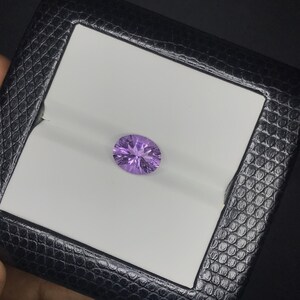 May include: A light purple oval-shaped gemstone with facets, set against a white background.