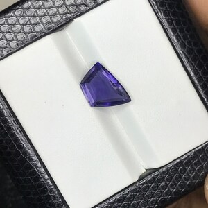 May include: A deep purple gemstone with a triangular shape, set in a white display box.