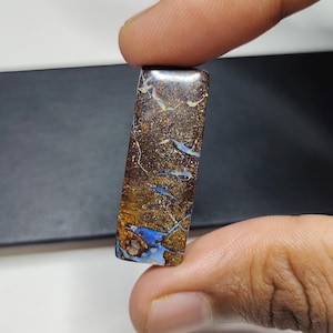 Może przedstawiać: Prostokątny, brązowo-niebieski opal szlachetny z unikalnym wzorem żyłek i zawirowań. Opal jest osadzony w przezroczystej, gładkiej i wypolerowanej oprawie.