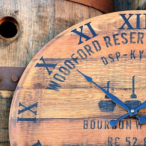 May include: A wooden clock face with black Roman numeral hour markers and black hands. The clock face is made from a repurposed bourbon barrel and features the text "WOODFORD RESERVE DIST DSP-KY-52 BOURBON WHISKEY RC 52.8 G".