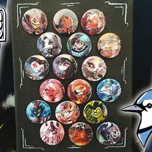 Può includere: Una collezione di 18 spille rotonde con diversi personaggi della serie animata "Hazbin Hotel". Le spille sono disposte in una griglia su uno sfondo nero. I personaggi includono: Lucifer, Charlie, Vaggie, Angel Dust, Alastor, e altri.