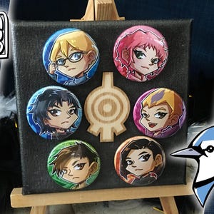 Könnte beinhalten: Eine schwarze Leinwand mit sechs runden Knöpfen, die Comicfiguren zeigen. Die Knöpfe sind in einem Kreis um ein Holzschild angeordnet. Das Schild ist ein stilisiertes Ziel mit einem Kreis in der Mitte und vier Armen, die nach außen ragen.