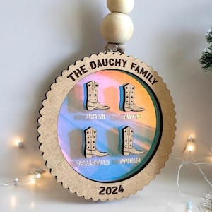 Op de afbeelding: Een houten kerstversiering met een lichtbruine achtergrond en een kleurrijk, iriserend, draaiend patroon erin. De versiering heeft een gegolfde rand en de tekst "THE DAUCHY FAMILY" bovenaan. Binnenin de versiering zijn vier kleine, bruine cowboylaarzen met de namen "RYAN", "TAYLOR", "SEBASTIAN" en "OPHILIA" gedrukt onder elke laars. Het jaar "2024" is onderaan de versiering gedrukt.