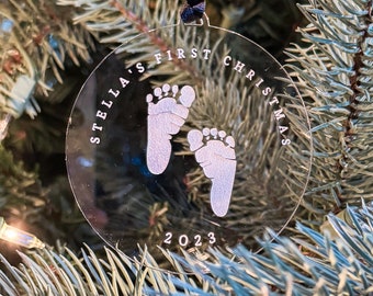 以下が含まれることがあります： 「Stella's First Christmas 2023」の文字と、2つの白い赤ちゃんの手形が描かれた、透明な丸いクリスマスオーナメントです。