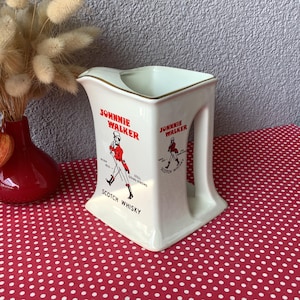 Op de afbeelding: Crèmekleurige keramische kan met het "Johnnie Walker"-logo en een figuur in rood. De kan heeft een vierkante basis en een handvat. De tekst "Scotch Whisky" staat onder het logo. Een rode vaas met droogbloemen staat op de achtergrond.