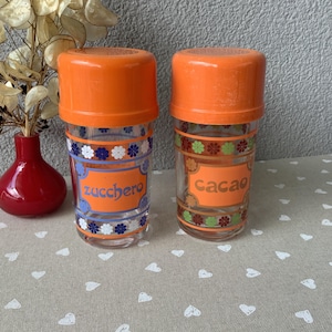 May include: Two vintage glass spice jars with orange lids. One jar reads "zucchero" in blue, the other "cacao" in orange. Both jars have decorative floral patterns in blue, white, green, and orange. A red vase and dried flowers are in the background.