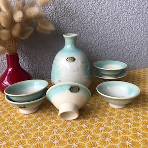 May include: A set of ceramic tableware featuring a red vase with dried flowers, a light blue and white sake bottle, and several small bowls. The bowls and bottle have a light blue rim. The items are arranged on a yellow patterned tablecloth.