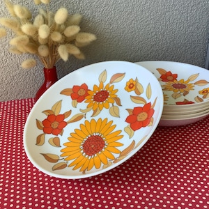 Könnte beinhalten: Ovale weiße Teller mit einem Retro-Blumenmuster mit orangefarbenen und gelben Sonnenblumen und Blättern. Ein Stapel Teller ist auf einer roten Oberfläche mit Polka-Dots zu sehen. Eine rote Vase mit Trockenblumen befindet sich im Hintergrund.