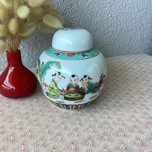 May include: A white ceramic ginger jar with a lid, featuring a hand-painted scene of children. The jar has a teal and red rim and a purple and gold base. A small red vase with dried flowers is next to the jar.