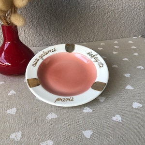 May include: A white ceramic ashtray with a pink interior and gold accents. The ashtray has the words "aux galeries lafayette paris" in gold lettering. A red vase with dried flowers is in the background.