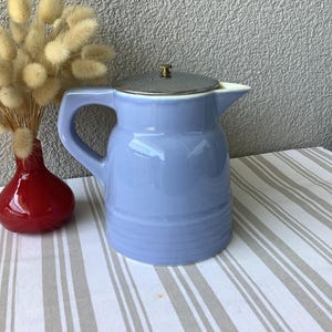 Peut inclure: Une cruche en céramique bleu clair avec un couvercle argenté et un petit bouton en laiton. La cruche a une anse et un bec verseur. Un vase rouge avec des fleurs séchées et des accents orange est à gauche.