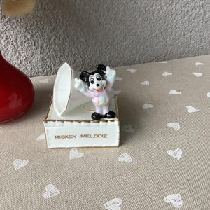 May include: A white porcelain Mickey Mouse figurine with black ears and shoes, and a pink and blue outfit, stands atop a white box with gold trim and the words "MICKEY MELODIE". A white, curved object is behind the figurine.