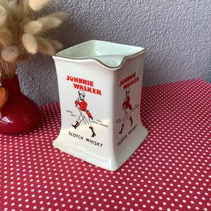K&ouml;nnte beinhalten: Vintage wei&szlig;e Keramikkrug mit Johnnie Walker-Logo, mit einer rot gekleideten Figur. Der Krug hat eine quadratische Form mit goldenem Rand und den Worten "Scotch Whisky". Eine rote Vase und Trockenblumen sind im Hintergrund.