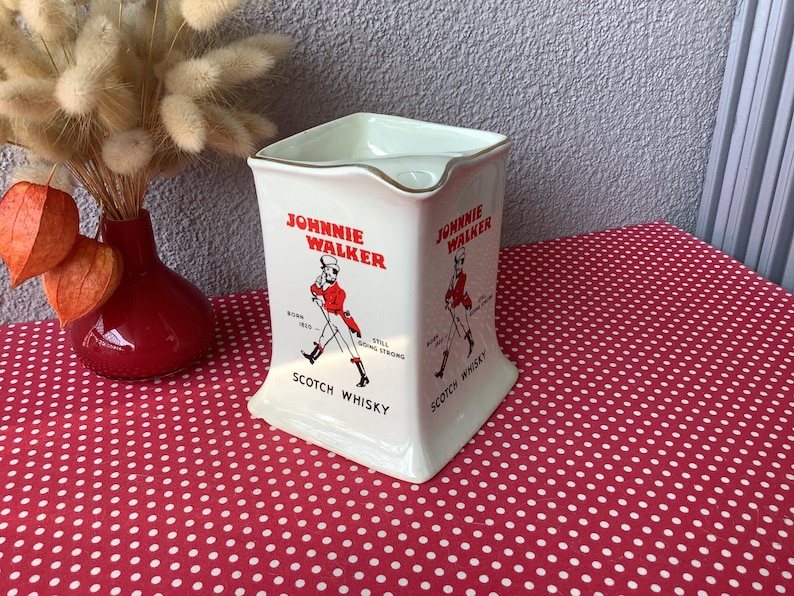 K&ouml;nnte beinhalten: Vintage wei&szlig;e Keramikkrug mit Johnnie Walker-Logo, mit einer rot gekleideten Figur. Der Krug hat eine quadratische Form mit goldenem Rand und den Worten "Scotch Whisky". Eine rote Vase und Trockenblumen sind im Hintergrund.