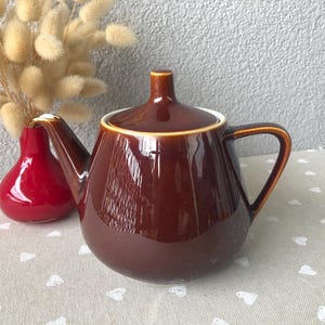 Può includere: Una teiera in ceramica marrone con coperchio, manico curvo e beccuccio. La teiera è accanto a un piccolo vaso rosso e a un mazzo di fiori secchi. La teiera ha una finitura lucida e un bordo marrone chiaro.