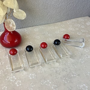 Peut inclure: Six supports en verre transparent avec des ornements ronds rouges et noirs sur le dessus. Les supports sont disposés en rangée sur une surface blanche à motif floral.
