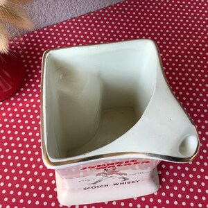 K&ouml;nnte beinhalten: Ein Vintage-Aschenbecher aus wei&szlig;er Keramik mit goldenem Rand und dem Johnnie Walker Scotch Whisky-Logo. Der Aschenbecher hat eine einzigartige Form mit einer gebogenen Seite und einer kleinen Ablage f&uuml;r eine Zigarette.