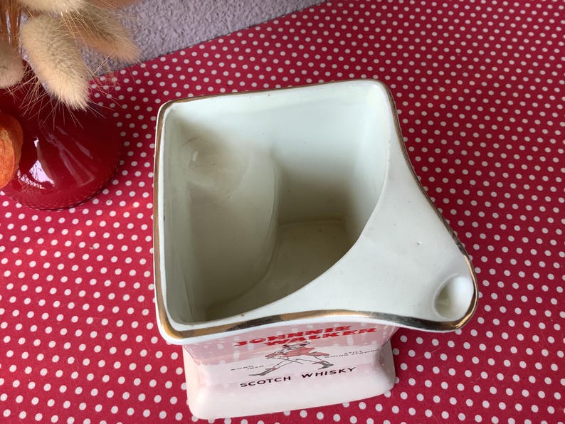 K&ouml;nnte beinhalten: Ein Vintage-Aschenbecher aus wei&szlig;er Keramik mit goldenem Rand und dem Johnnie Walker Scotch Whisky-Logo. Der Aschenbecher hat eine einzigartige Form mit einer gebogenen Seite und einer kleinen Ablage f&uuml;r eine Zigarette.