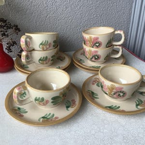 Könnte beinhalten: Ein Set mit sechs Teetassen und Untertassen mit floralem Muster. Die Tassen und Untertassen sind weiß mit einem rosa und grünen Blumendesign.