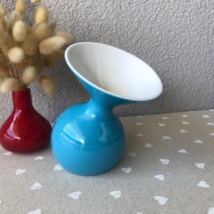 May include: A vibrant turquoise ceramic vase with a white interior, next to a small red vase. The turquoise vase has a unique, curved design. The red vase holds dried, fluffy beige flowers. Both vases sit on a patterned surface.