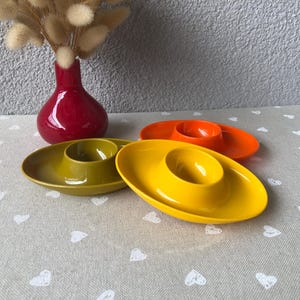 Op de afbeelding: Drie eierdopjes in een ovale vorm in de kleuren geel, olijfgroen en oranje. Elk eierdopje heeft een gladde, glanzende afwerking met een centrale ronde inkeping die is ontworpen om een enkel ei vast te houden, gerangschikt in een groep.