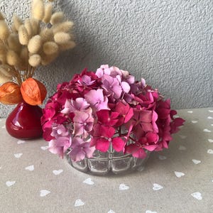 Può includere: Una ciotola di vetro trasparente piena di ortensie rosa e viola. Accanto, un piccolo vaso rosso contiene fiori secchi e lanterne cinesi arancioni. La composizione è su una tovaglia beige con motivi a cuore bianchi.