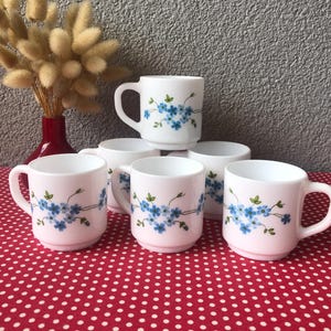 May include: A set of five white milk glass mugs featuring a delicate pattern of small blue flowers with green leaves and stems. Each mug has a rounded handle and a smooth finish, arranged in a cluster on a red surface with white polka dots.